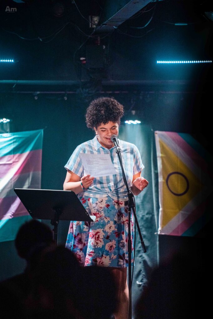 A photo of Laura reading a poem on a stage, in front of a trans flag and an Intersex-Inclusive Progress Pride flag.