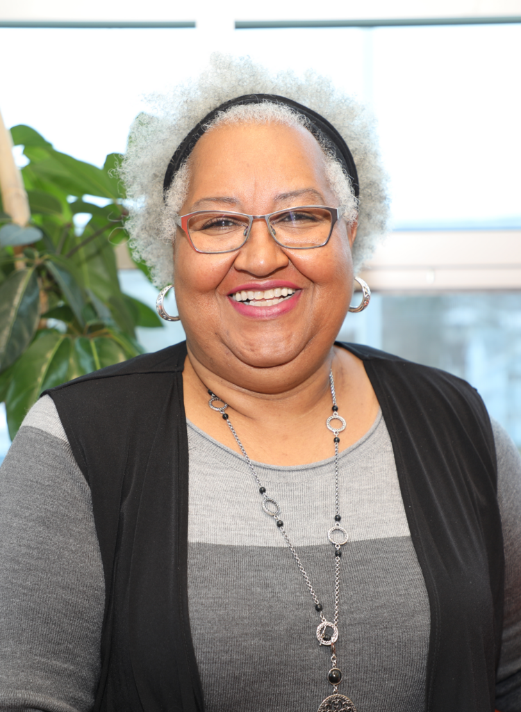 Photo of Shelley Fashan, a Black woman with curly grey hair. She is wearing glasses, a black headband and a black vest over a grey sweater.